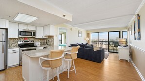 A modern kitchen with a central island, bar stools, a dining area with a sofa, and a balcony with a view. - Summer Beach 608 by Seaside Vacations & Sales (Ocean City)