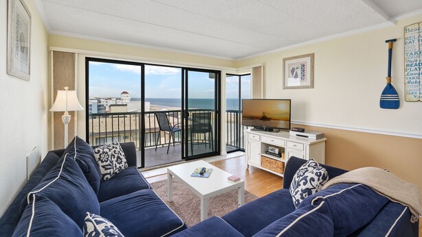 A living room with a sofa set, a TV, and a view of the beach. - Summer Beach 608 by Seaside Vacations & Sales (Ocean City)