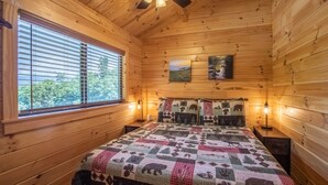 A wooden cabin bedroom with a bed, two pillows, a ceiling fan, and a window with blinds.