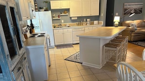 Una cocina moderna con armarios blancos, una isla central con taburetes, una zona de comedor con mesa y sillas, y un salón con un sofá y una lámpara.