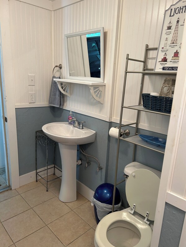 Un baño con lavamanos pedestal, inodoro, cubo de basura azul, estante metálico y espejo instalado en la pared.