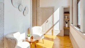 Una habitación moderna con piso de madera, paredes blancas y una puerta que da a otra estancia. Hay dos sillas blancas y una pequeña mesa con una planta.