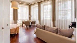 A spacious living room with a sofa, a dining table with chairs, and large windows with curtains. - Traça Apartments by DA'HOME - Studio Deluxe #1 (Porto)