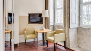 A hotel room with a large window, a flat-screen TV mounted on the wall, two armchairs, a small round table, and a lamp. - Traça Apartments by DA'HOME - Studio #2 (Porto)