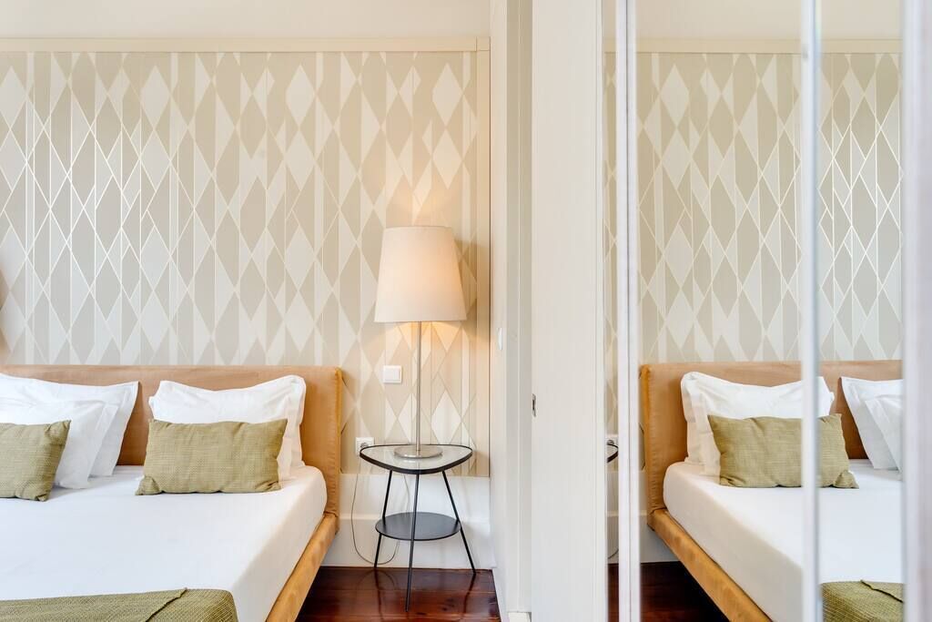 Habitación de hotel con dos camas, una mesita redonda pequeña con lámpara y pared con diseño geométrico.