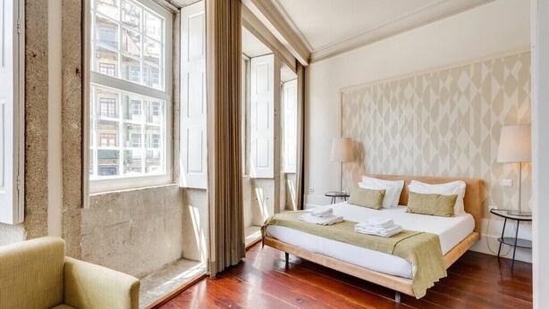 A hotel room with a large bed, a chair, and a view of buildings through the windows. - Traça Apartments by DA'HOME - Studio #2 (Porto)