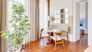 Una camera d'albergo moderna con pavimento in legno, un tavolo rotondo con due sedie, una pianta in vaso e un'ampia finestra con tende leggere.