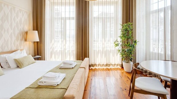 Una camera d'albergo con un letto, un comodino, una lampada, una pianta in vaso e un'area ristorazione con tavolo e sedie.