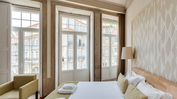 Habitació d'hotel amb un llit, una cadira i vista a edificis a través de grans finestres.