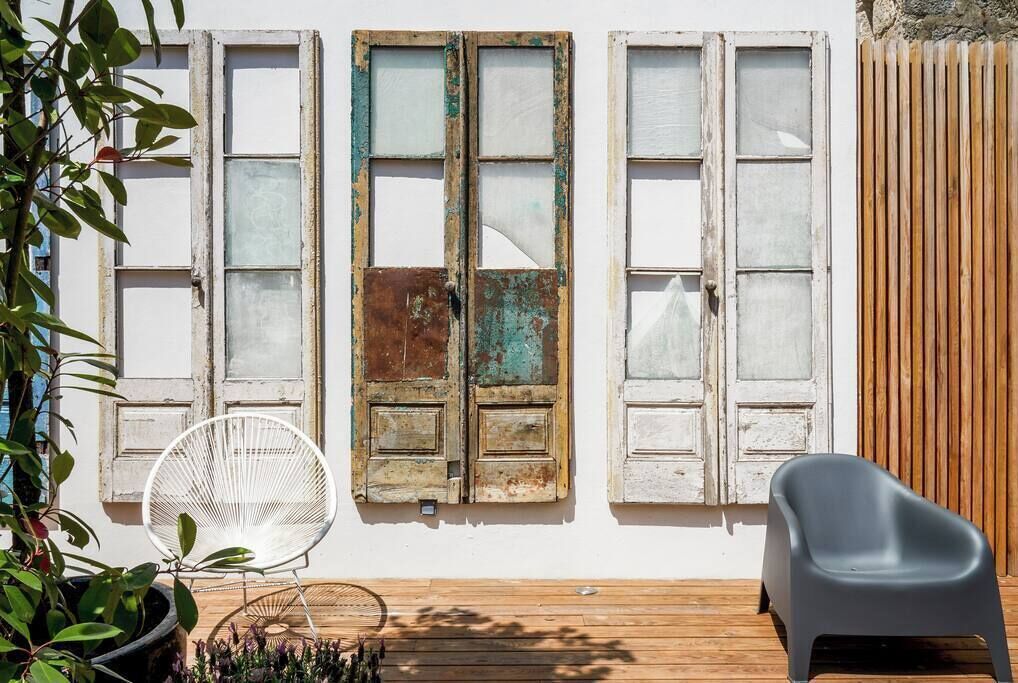A wooden deck with two white chairs, a potted plant, and a wall with three weathered windows.
