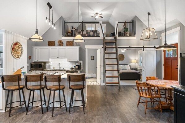 Una cocina amplia con armarios blancos, una isla central con taburetes y una zona de comedor con mesa y sillas de madera. Se ve un dormitorio entrepiso sobre la cocina, y una escalera conduce a él.
