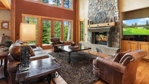 Un salon spacieux avec une cheminée en pierre, des canapés en cuir et une grande fenêtre donnant sur une forêt.