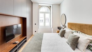 A modern bedroom with a large bed, a TV mounted on the wall, and a window with a view of buildings.