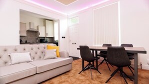 Un salon moderne avec un canapé gris, une table à manger entourée de quatre chaises et une cuisine visible à travers une porte ouverte.