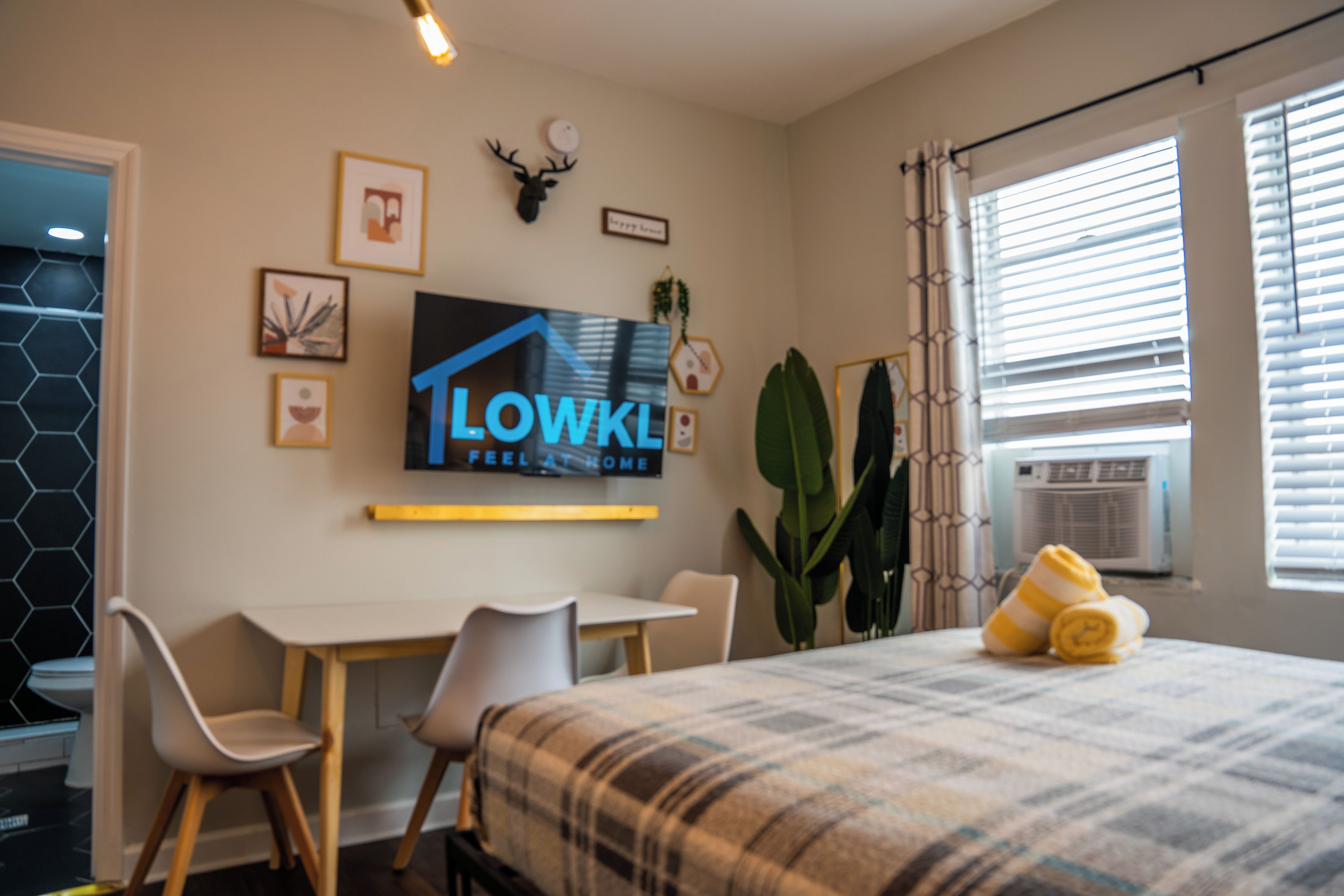A hotel room with a bed, a desk, two chairs, a television mounted on the wall, and a large window with blinds.