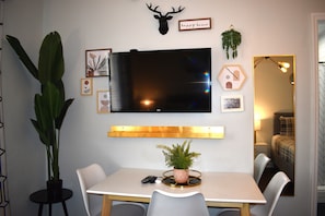 Habitació amb televisió penjada, taula de dinar amb cadires, una planta en test i un llit amb llençol escocès.