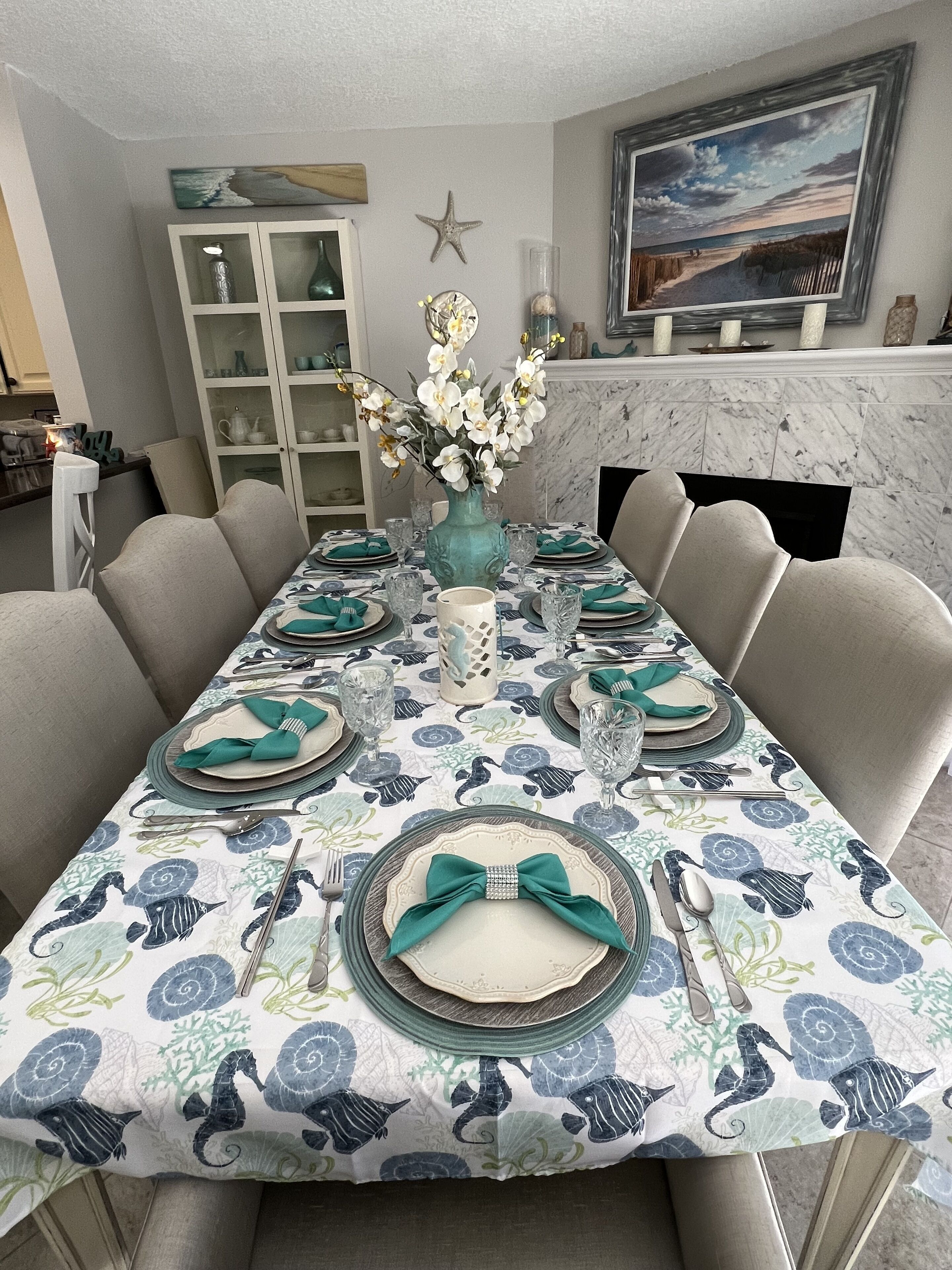A dining table set with a floral tablecloth, a vase of flowers, and a framed picture on the wall.