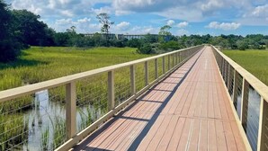 A wooden boardwalk over a water body with a metal railing, surrounded by greenery and a clear sky.
