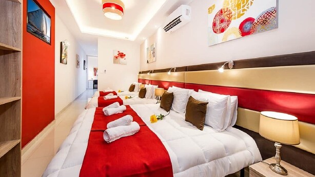A hotel room with a long bed, red and white bedding, a headboard with a red and gold design, a bedside table with a lamp, and artwork on the walls. - Orange 2: Studio Apartment in St. Julian's (Swieqi)