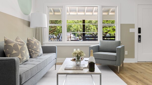 A modern living room with a grey sofa, a grey armchair, a glass coffee table, and a large window with a view of trees and buildings. - Steps From Park Circle | Mavis at the Quad (North Charleston)