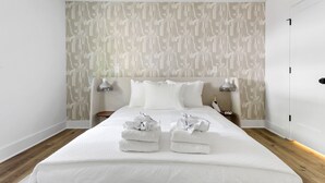 A modern hotel room with a large bed, white linens, and a patterned wallpaper. There are two bedside tables with lamps and a small nightstand with a lamp. - Steps From Park Circle | Mavis at the Quad (North Charleston)