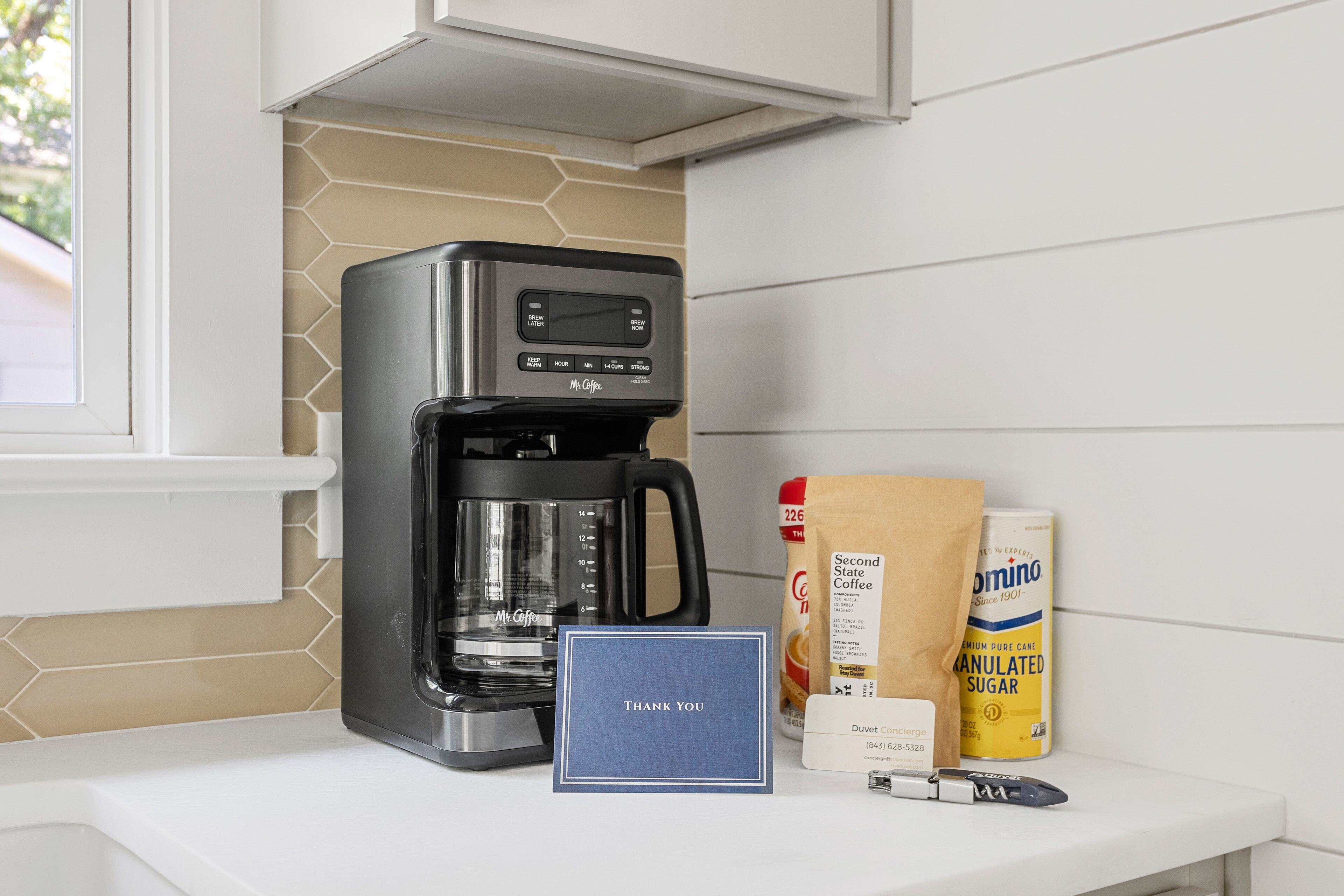A coffee maker, a bag of coffee, a bag of sugar, a card, and a can of coffee on a countertop.