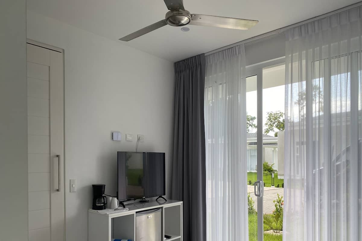 A hotel room with a bed, a TV on a stand, a ceiling fan, and a view of the outdoors through sheer curtains.