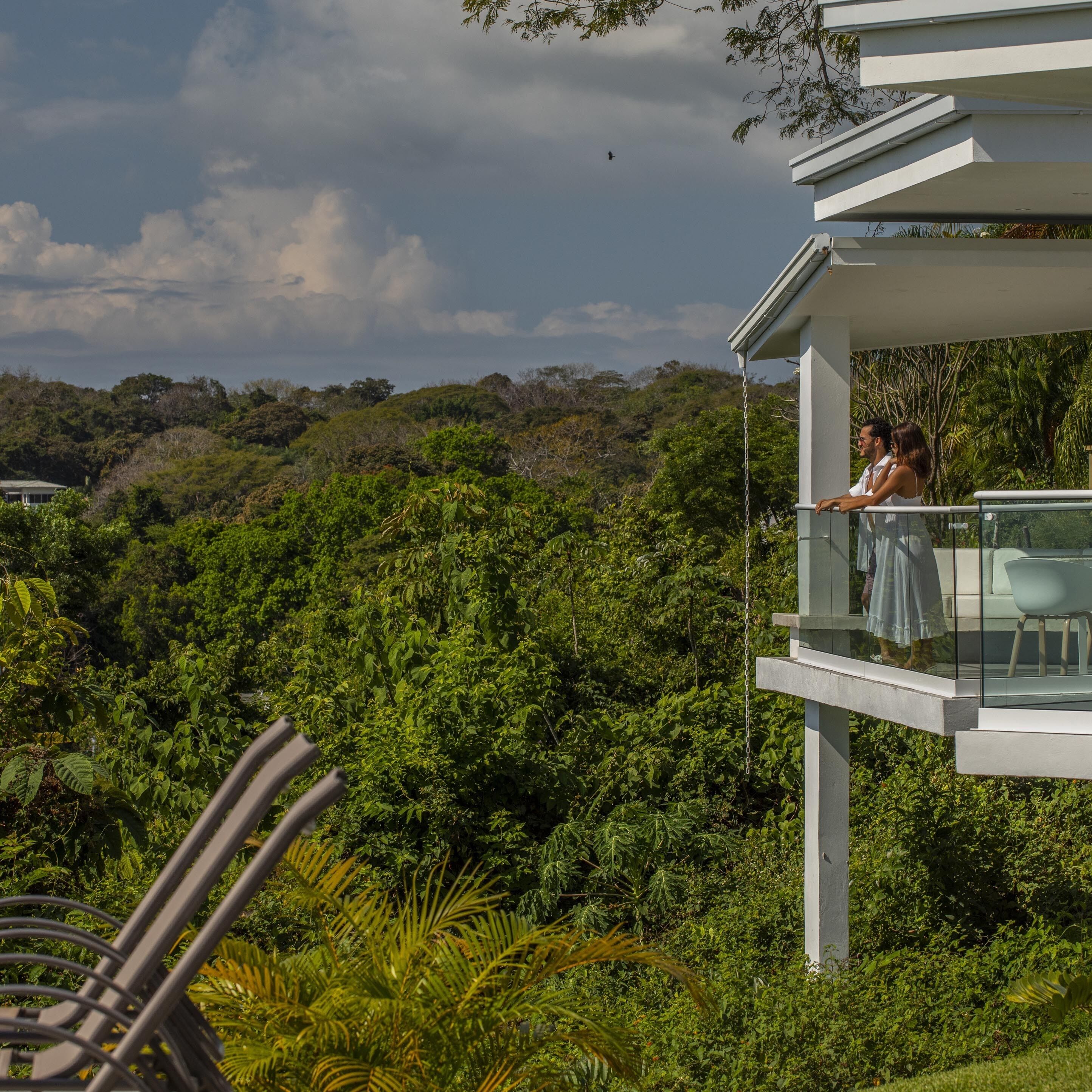 A balcony with a glass railing overlooking lush greenery and a distant view of a building.
