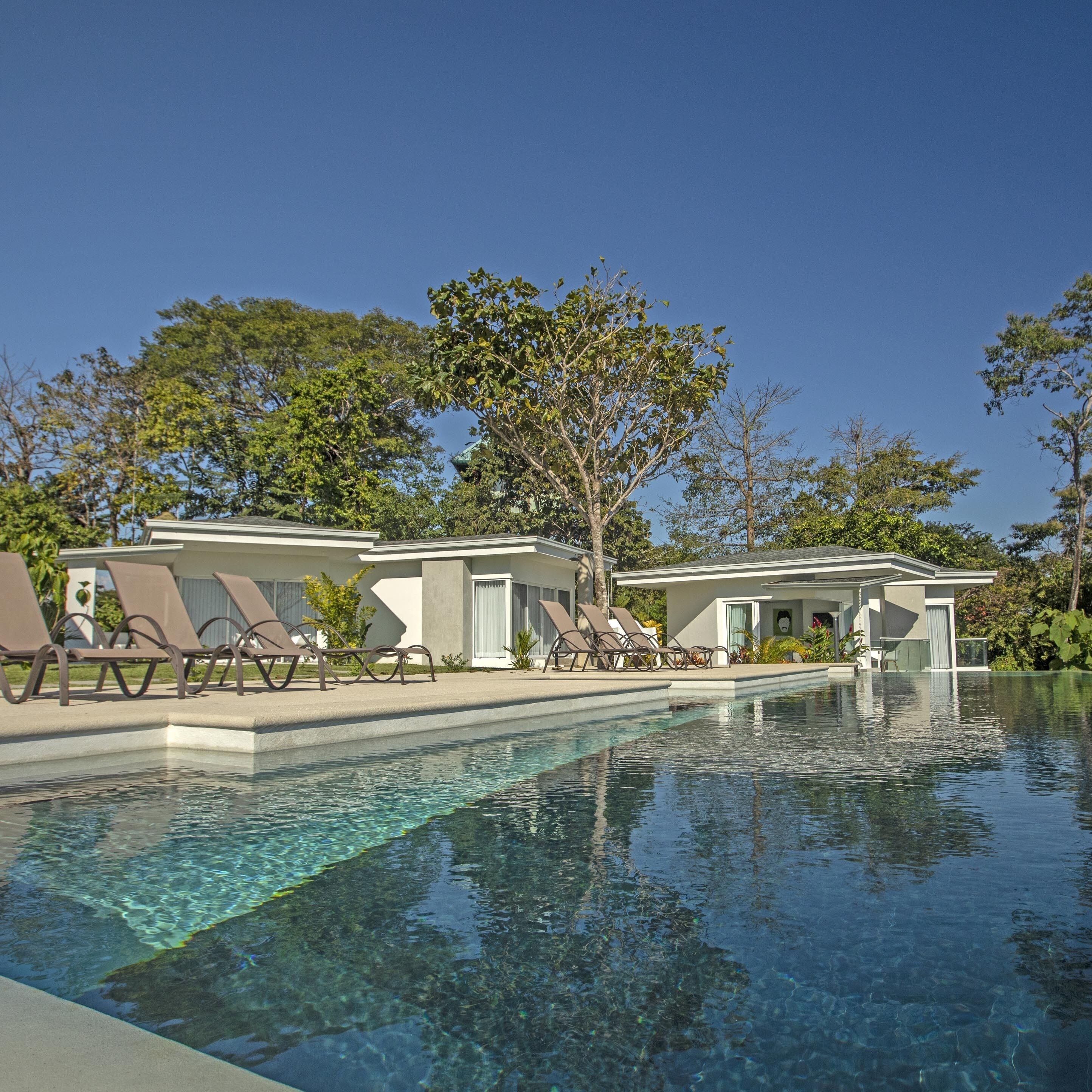 Una piscina con agua cristalina, rodeada de sillas de descanso y un edificio moderno con un patio cubierto.