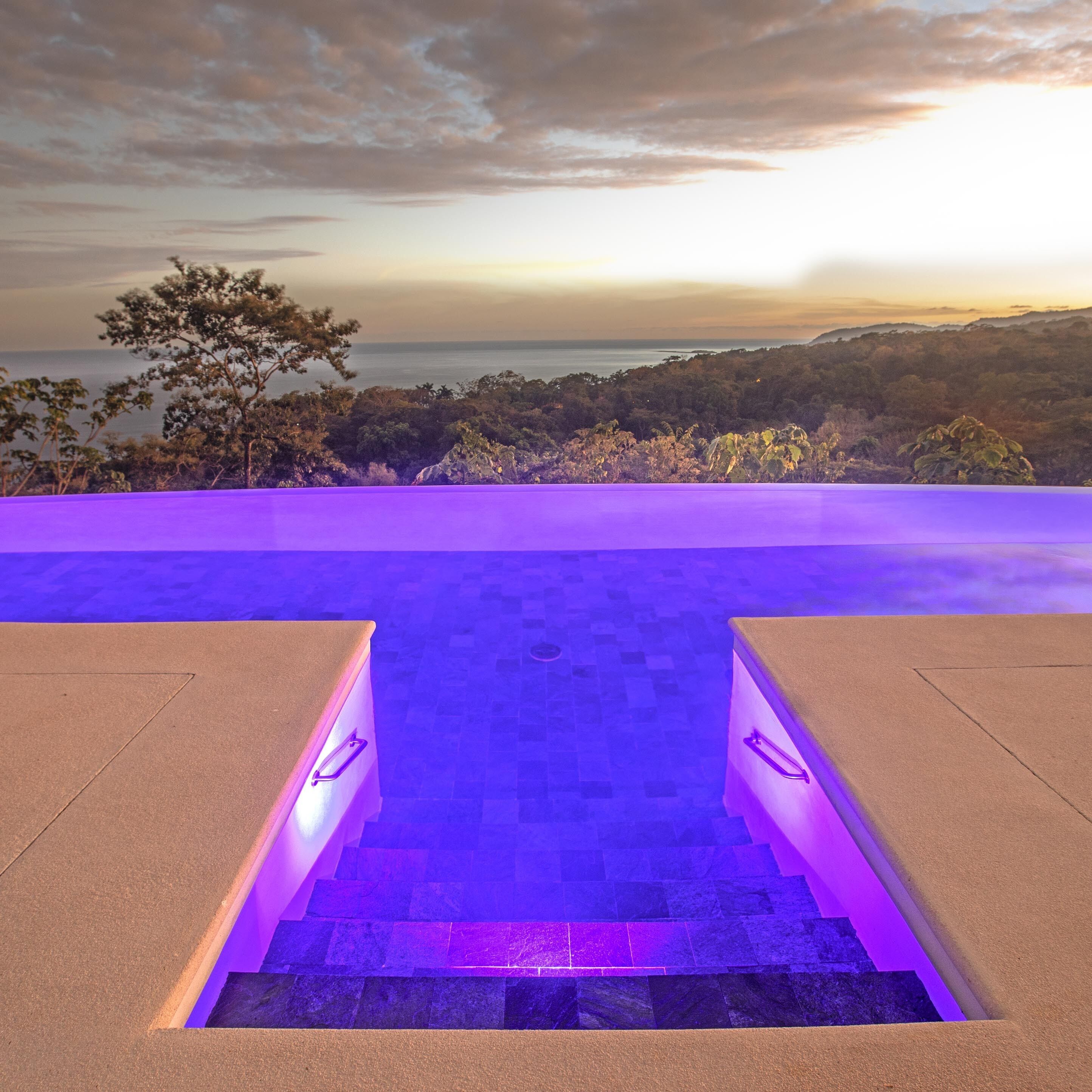 A pool area with a view of the ocean at sunset, surrounded by trees.
