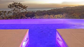 A pool area with a view of the ocean at sunset, surrounded by trees.