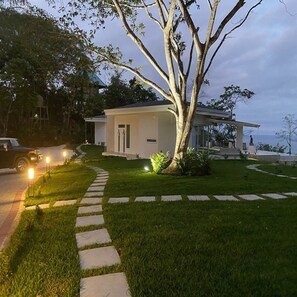 A modern house with a paved driveway, a well-maintained lawn, and a tree in the foreground. - Adeluna Luxury Ocean View Villas (Cóbano)