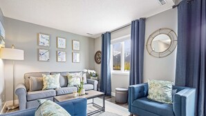 Un salon moderne avec un canapé gris, des fauteuils bleus et une table basse. Les murs sont ornés de tableaux encadrés, il y a un miroir rond et une fenêtre avec des rideaux.