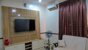 A living room with a flat-screen TV mounted on a wooden wall, a white sofa, a small coffee table, and a fan.