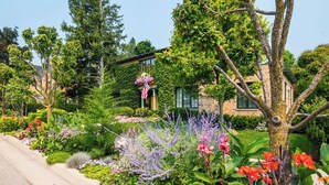 A well-maintained garden with various plants and flowers, a stone building with a green roof, and an American flag. - Riversbye: Historic luxury riverfront retreat (Boyne City)