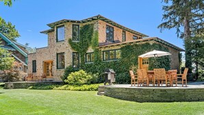 A two-story house with a stone facade, a covered patio with outdoor furniture, and a well-maintained lawn. - Riversbye: Historic luxury riverfront retreat (Boyne City)