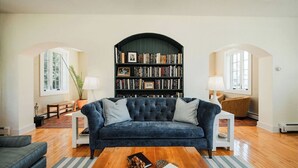 A living room with a dark blue sofa, a wooden coffee table, a bookshelf, and a lamp. - Riversbye: Historic luxury riverfront retreat (Boyne City)