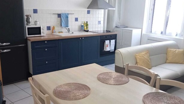 A compact kitchen with a microwave, sink, and stove. There's a dining table with placemats and a sofa in the adjacent living area. - Joli Appartement Avec 2 Chambres (Périgueux)