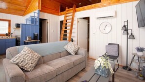 Un salón acogedor con techo de madera, un sofá con cojines estampados, una escalera, un televisor y una pequeña mesa con una planta.