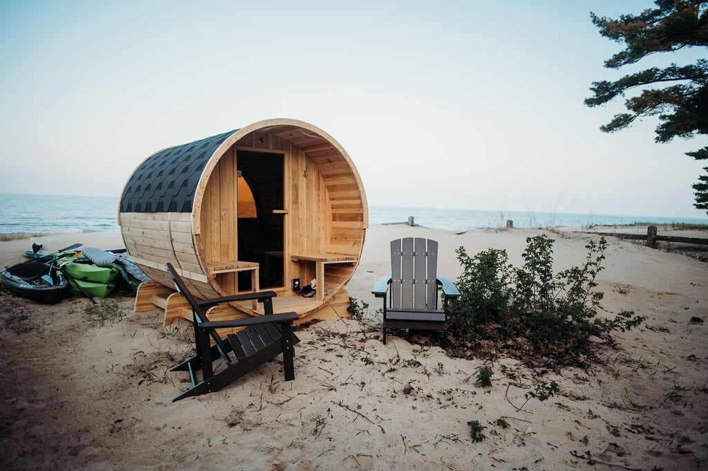 A wooden barrel-shaped structure on a beach with a chair and a bench inside.
