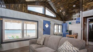 Una sala de estar acogedora con un sofá gris, cojines estampados y amplias ventanas con persianas.