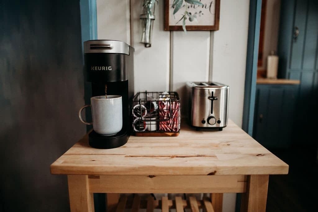 Een houten tafel met een Keurig koffiezetapparaat, een witte mok, een mand met een rood-wit patroon, en een zilveren broodrooster.