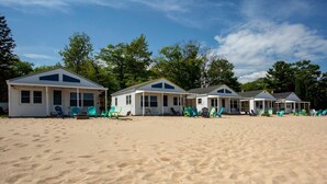 A row of beach cottages with multiple windows and doors, arranged along a sandy beach with green chairs outside. - Endless Adventures at Lake Huron Cottage 4 (Greenbush)
