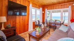A cozy cabin interior with a wooden interior, a TV mounted on the wall, a sofa, a coffee table with books and a plant, and a dining area with chairs and a table.
