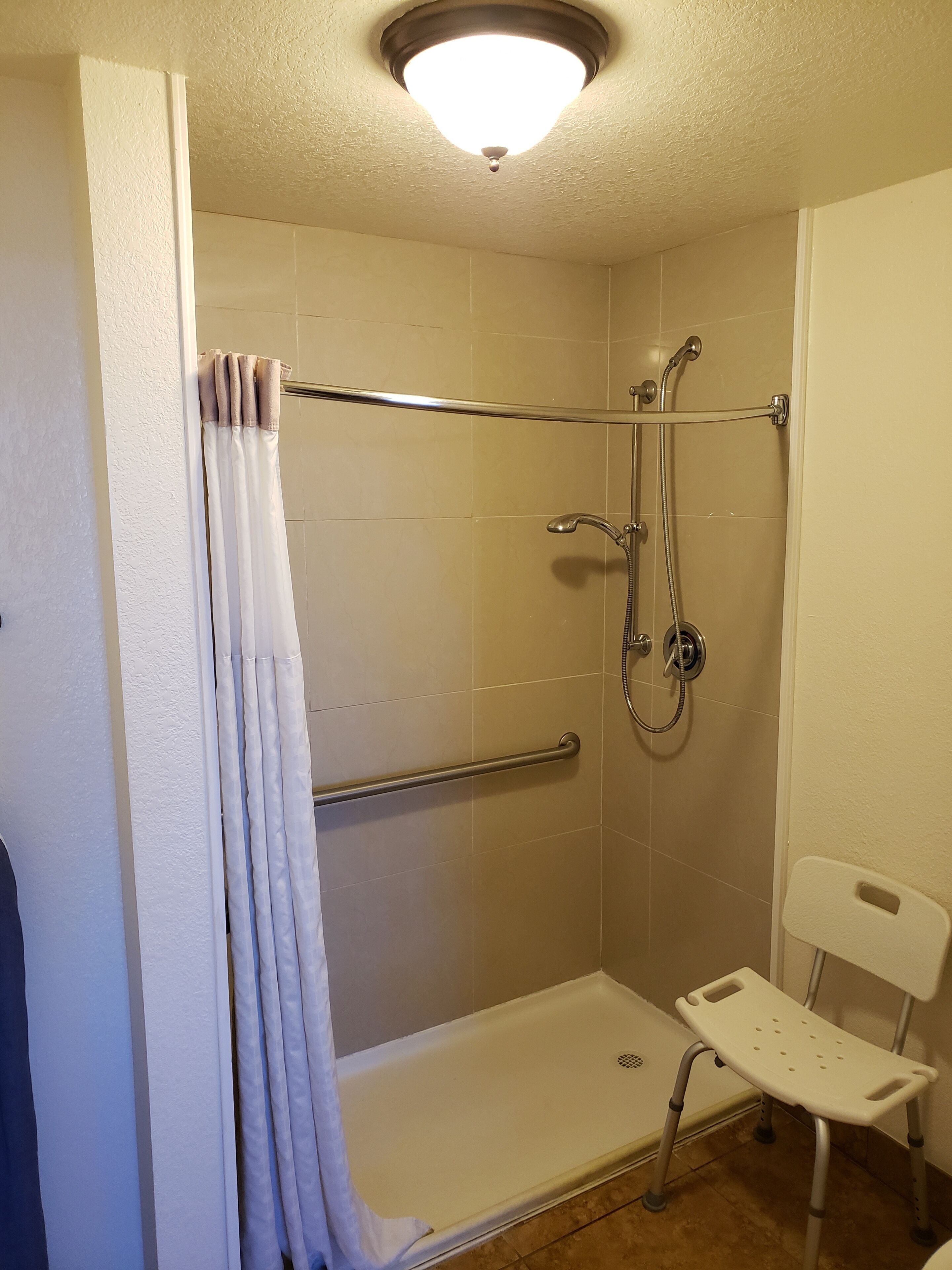 Un espacio de ducha con puerta de cristal, barra de apoyo, ducha y una pequeña silla blanca.
