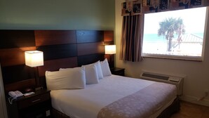 A hotel room with a large bed, two bedside lamps, a telephone, and a window with a view of palm trees and the sea. - Oceanfront Inn and Suites (Ormond Beach)