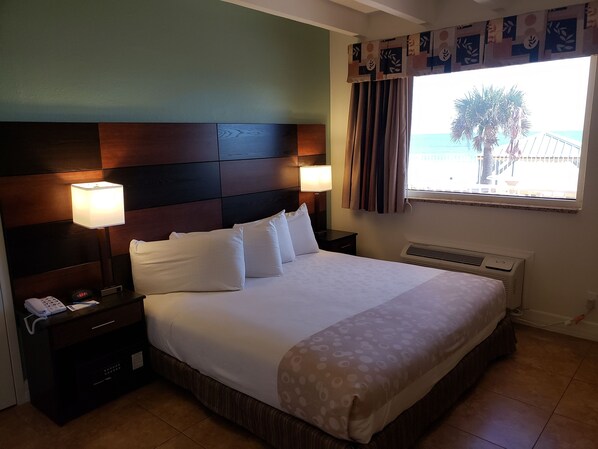 Ein Hotelzimmer mit einem großen Bett, zwei Nachttischlampen, einem Telefon und einem Fenster mit Blick auf Palmen und das Meer.
