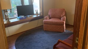 A living room with a television on a wooden shelf, a pink armchair, a blue rug, and a wooden door. - Walk to Beach, shops, restaurants and much more! (Long Beach)