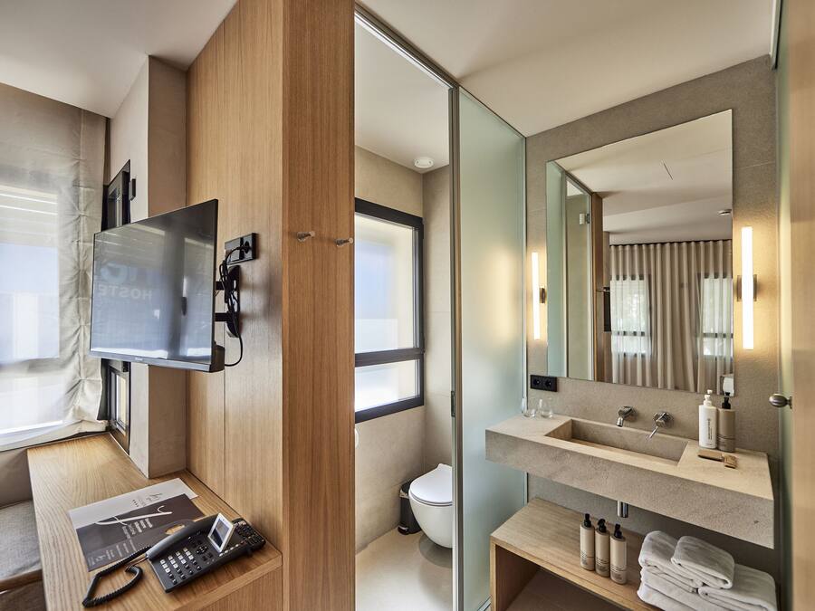 Un baño moderno con un espejo grande, un lavamanos y un inodoro. Se aprecia panelado de madera en una pared y una ventana que deja entrar luz natural.
