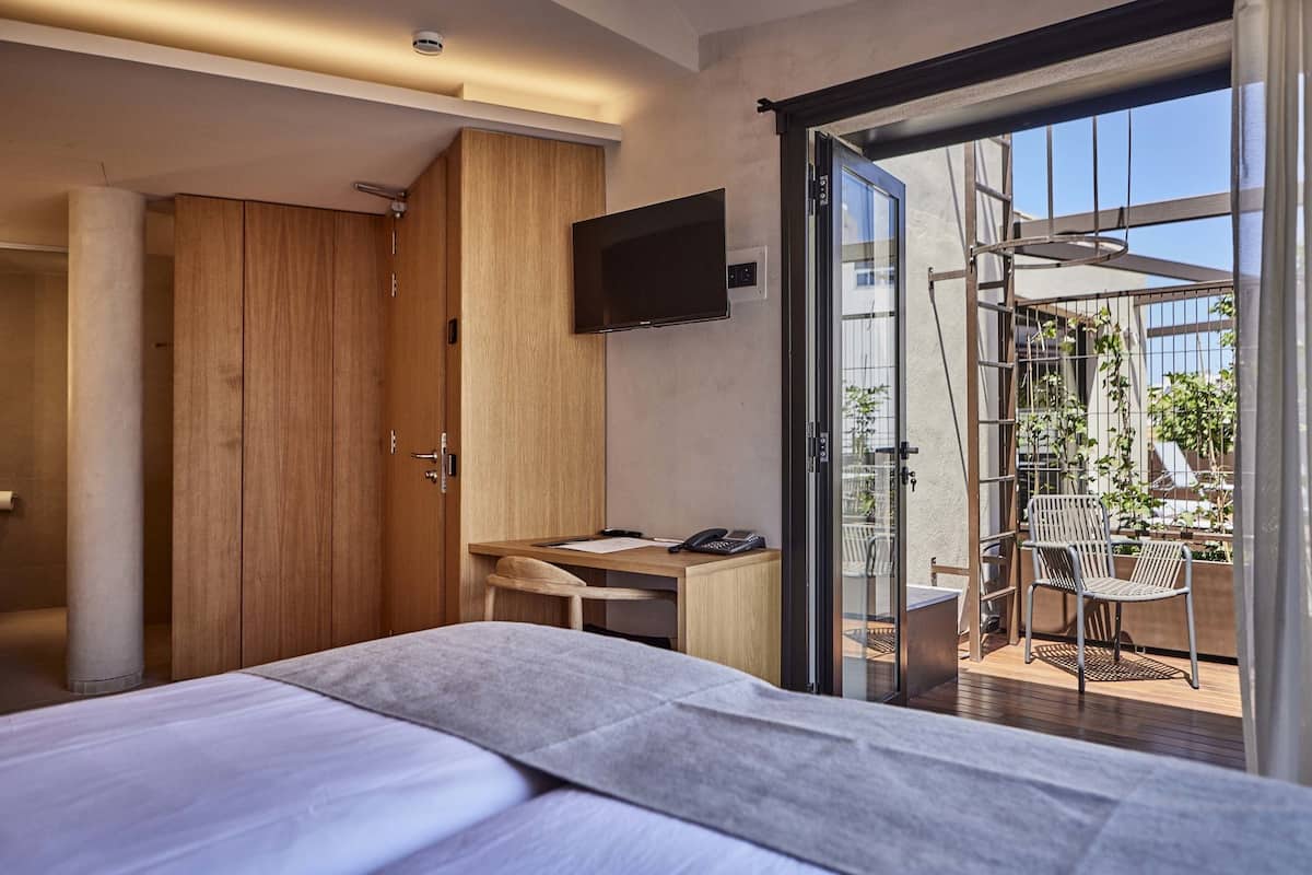 Une chambre d’hôtel moderne, équipée d’un lit, d’un bureau et d’une télévision. Elle dispose d’un balcon avec des chaises et des plantes.